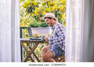 Attractive young man laughs happily while talking to his laptop on a tropical balcony. He holds his coffee in one hand and wears a hat.