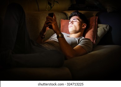 Attractive Young Man Lying On Sofa At Night And Illuminated By Light From Screen Of Cell Phone At Home In Living Room