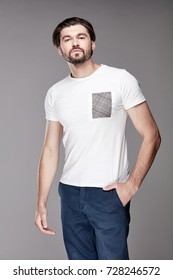 Attractive Young Man Model With Beard And Dark Hair Posing In White Tshirt With Grey Pocket And Dark Blue Trousers In Light Studio