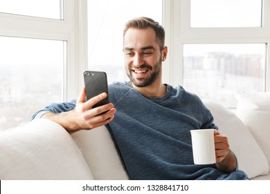 Attractive young man relaxing on a couch at home, using mobile phone, drinking coffee