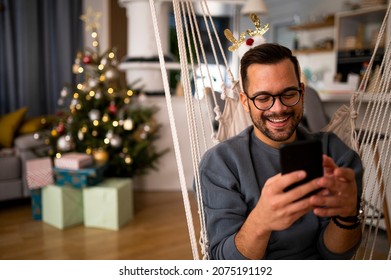 Attractive young man sitting and holding his smartphone video chatting at a christmas party in the apartment in front of a tree and presents. - Powered by Shutterstock