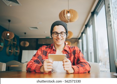 Attractive Young Man Is Sitting In Office And Drinking Tea. He Is Holding A Mobile Phone And Watching Video Or Reading Interesting Article. Handsome Guy Is Smiling