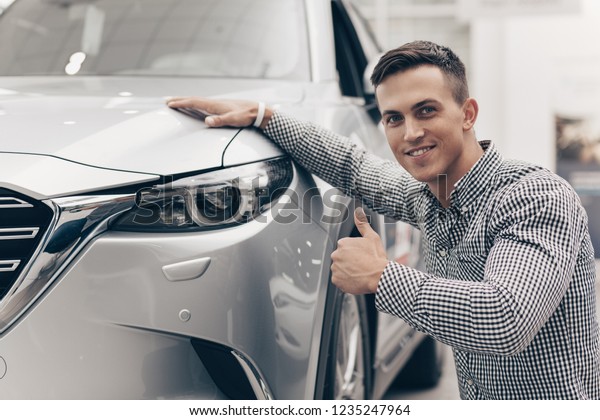 Attractive young man\
smiling, showing thumbs up, posing near his new automobile at car\
dealership. Happy male customer buying car. Ownership rental\
service concept