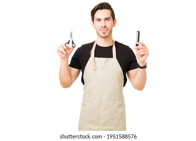 Attractive Young Man Smiling While Making Eye Contact And Holding A Comb With Some Scissors. Male Hair Stylist Working At The Barber Shop