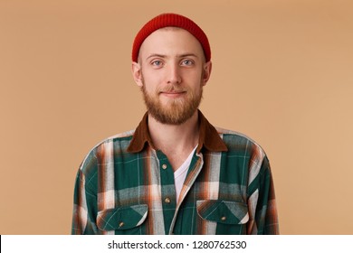 Attractive Young Man In Studio Looking At Camera. Portrait Of A Normal Guy With Broad In Red Hat And Checkered Shirt In Front Of Beige Background