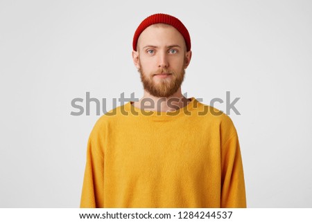 Attractive young man in studio looking at camera.Calm interesting bearded hipster guy in red hat with normal face expression,over white background. Man takes photo for passport at local studio
