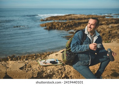 Attractive young man tourist sitting on a rock by sea, enjoying a hot beverage in thermos flask. Happy male hiker relaxing while drinking a cup of coffee during the hiking on the adventure travel. - Powered by Shutterstock