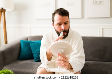 Attractive Young Man Wearing A Soft Robe And Trimming His Nose Hair With An Electric Trimmer