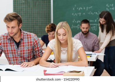 Attractive Young Man And Woman Studying Together At Their Desks In A College Or University Classroom With Other Students And A Blackboard Behind Them