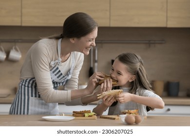 Attractive young mother feeds her cute preschooler daughter with fresh prepared meat or vegan sandwiches, express care, feel love, spend time together, stand in cozy kitchen at home. Cookery, cuisine