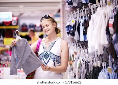 Attractive Young Mother Shopping For Childrens Clothes In A Retail Clothing Store Viewing Items On A Rack