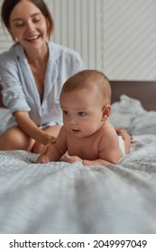 Attractive Young Mother Sitting On Bed Helping Young Naked Baby To Lay On Tummy. Tummy Time In Bed For Cute Infant Baby. Indoor Portrait Mother And Child On Family Bed.