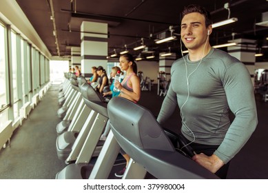 Attractive Young Muscular Man In Headphones Running On A Treadmill In Gym  And Smiling