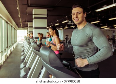 Attractive Young Muscular Man In Headphones Running On A Treadmill In Gym, Looking At Camera And Smiling