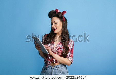 Attractive young pinup woman in retro shirt using tablet pc over blue studio background. Pretty lady in vintage style outfit shopping online or surfing web, watching video