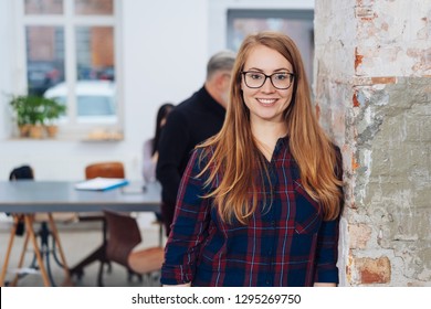 Attractive young redhead businesswoman standing in an office smiling at the camera as she leans against on old concrete pillar