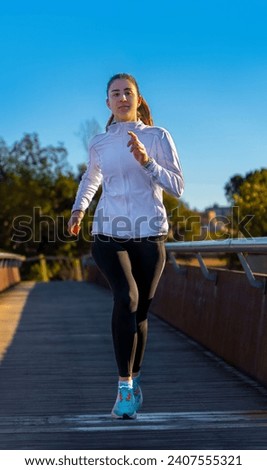 Attractive young runner girl, dressed in black tights and white sports jacket, running training in the morning on a wooden floor bridge under the dawn light of a public park.