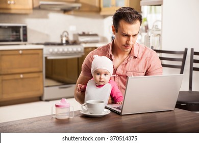 Attractive Young Single Dad Working On A Laptop Computer At Home While Taking Care Of His Baby Daughter