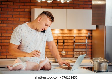 Attractive Young Single Dad Working On A Laptop Computer At Home While Taking Care Of His Baby Son