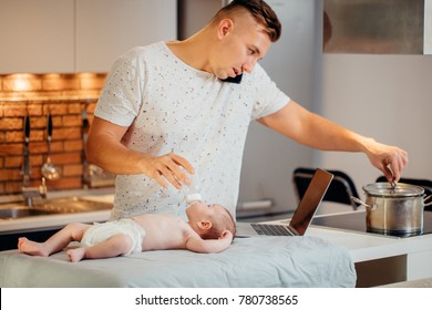 Attractive Young Single Dad Working On A Laptop Computer At Home While Taking Care Of His Baby Son