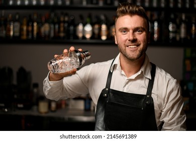 Attractive Young Smiling Male Bartender In Black Leather Apron Holding A Glass Shaker In His Hand. Blurred Shelves With Bottles In The Background