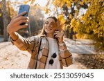 attractive young smiling woman walking in autumn park taking selfie pictures with leaf using smartphone, wearing checkered coat, happy mood, fashion style trend