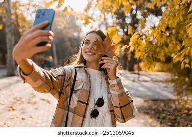 attractive young smiling woman walking in autumn park taking selfie pictures with leaf using smartphone, wearing checkered coat, happy mood, fashion style trend - Powered by Shutterstock