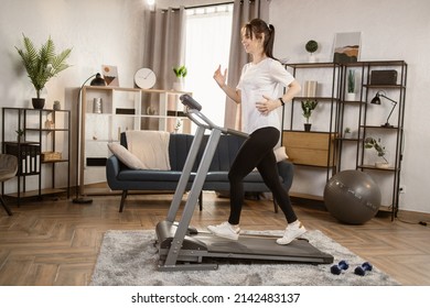 Atractiva joven mujer caucásica deportiva corriendo en cinta de correr. Haciendo ejercicio físico. Una mujer atlética y muscular ejercita activamente en el moderno apartamento. Las mujeres deportivas entrenan en casa. Vista lateral