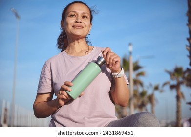 Attractive young sporty female, sportswoman, athlete holding a bottle with water, smiling, looking into distance, standing on the beach, resting after outdoor workout or morning jog. People and sport - Powered by Shutterstock