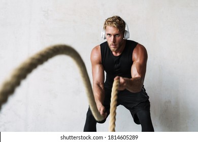 Attractive young sporty man in headphones exercising with a fitness rope over gray wall background, indoors - Powered by Shutterstock