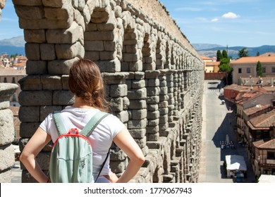 An Attractive Young Tourist Women With A Bag On Her Back Stares Fixedly At Te Famous Ancient Aqueduct Of Segovia.