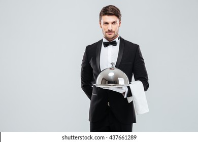 Attractive young waiter in tuxedo holding serving tray with metal cloche and napkin