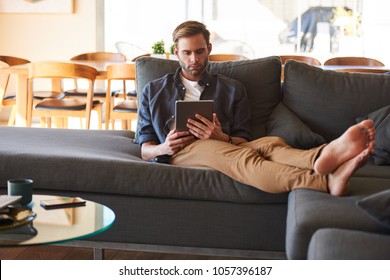 Attractive Young White Man Sitting Comfortably On His Great Couch With His Feet Up While Surfing The Internet On His Electronic Tablet To Keep Track Of The Latest News From Around The World.