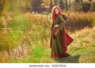 Attractive Young Woman Archer With Magnificent Long Red Hair In A Historical Celtic Dress Is Posing Near The Lake. Historical Reconstruction. 