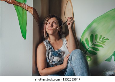 Attractive Young Woman Artist Looking Away Thoughtfully Holding Some Paintbrushes In Her Home Wearing An Old-fashioned Artist's Smock Which Is Covered With Paint Marks