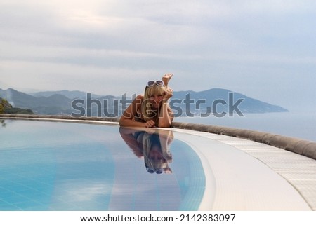 Attractive young woman in bikini enjoying a hot day, relaxing on the edge of the pool.The girl lies on her stomach, leaning on her elbows against the backdrop of mountains and the sea.Vacation concept