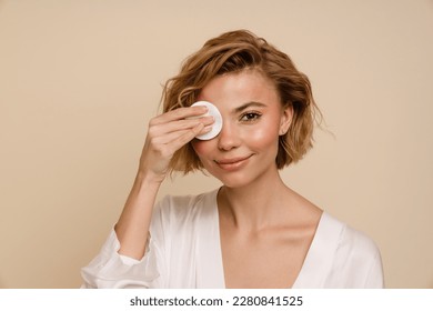 Attractive young woman with blonde hair covering her eye with cotton pad while standing isolated over beige studio wall - Shutterstock ID 2280841525