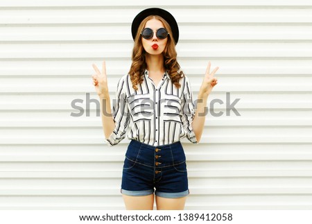 Attractive young woman blowing red lips sending sweet air kiss in black round hat, shorts, white striped shirt on white wall background