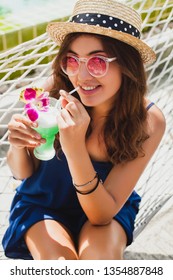 attractive young woman in blue dress and straw hat wearing pink sunglasses drinking alcohol cocktail on vacation sitting in hammock in summer style outfit, smiling happy in party mood