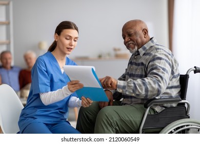 Attractive young woman in blue workwear nurse helping senior black man in wheelchair with questionnaire, african american pensioneer filling papers at nursing home, having assistance