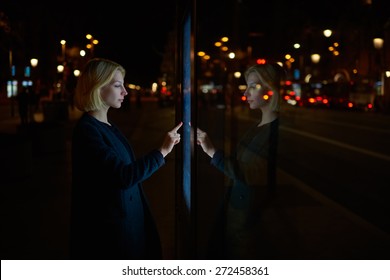 Attractive young woman consults on modern big timetable outdoors, female caucasian tourist touching information kiosk digital screen while standing at night city with beautiful lights on background