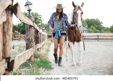 Attractive young woman cowgirl in hat smiling and walking with her horse in village