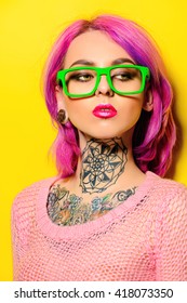Attractive young woman with crimson hair wearing bright green glasses posing over yellow background. Bright style, fashion. Optics style. Tattoo. Hair coloring.