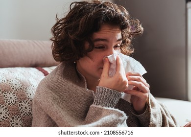 Attractive Young Woman With Curly Brown Hair, Wrapped In Warm Blanket, Sits On Comfortable Sofa At Home, Blowing Her Nose In Disposable Tissue. Allergy To Dust, Animal Dander, Cold, Or Viral Infection