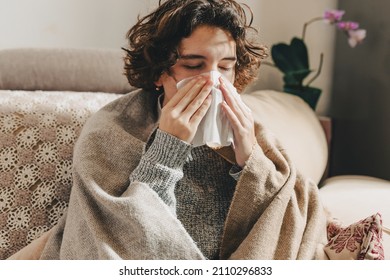 Attractive young woman with curly brown hair, wrapped in warm blanket, sits on comfortable sofa at home, blowing her nose in disposable tissue. Allergy to dust, animal dander, cold, or viral infection