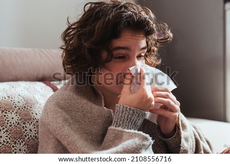 Attractive young woman with curly brown hair, wrapped in warm blanket, sits on comfortable sofa at home, blowing her nose in disposable tissue. Allergy to dust, animal dander, cold, or viral infection