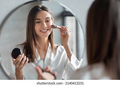 Attractive Young Woman Doing Daily Makeup While Standing Near Mirror In Bathroom, Happy Beautiful Female Putting Blush On Cheeks While Getting Ready At Home, Selective Focus On Reflection - Shutterstock ID 2140413231