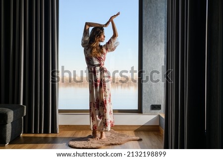 Attractive young woman dressed in a dressing gown dances in front of a huge window in the room. Sunny morning. The girl stands on tiptoe and looks away.