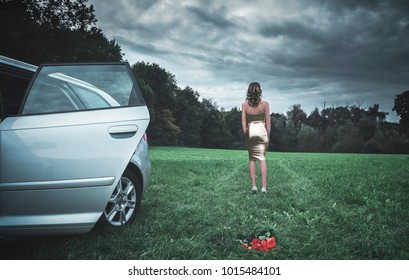 Attractive Young Woman Dressed In A Golden Latex Dress, Getting Away From A Car And A Bouquet Of Roses Lying Down On The Grass, On A Gloomy Day.