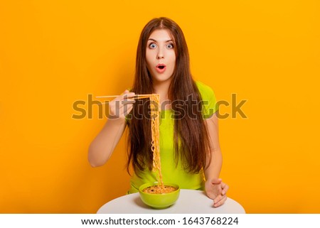 Attractive, young woman eating instant noodles with chopsticks from a green bowl, sitting at a white table on a yellow background.